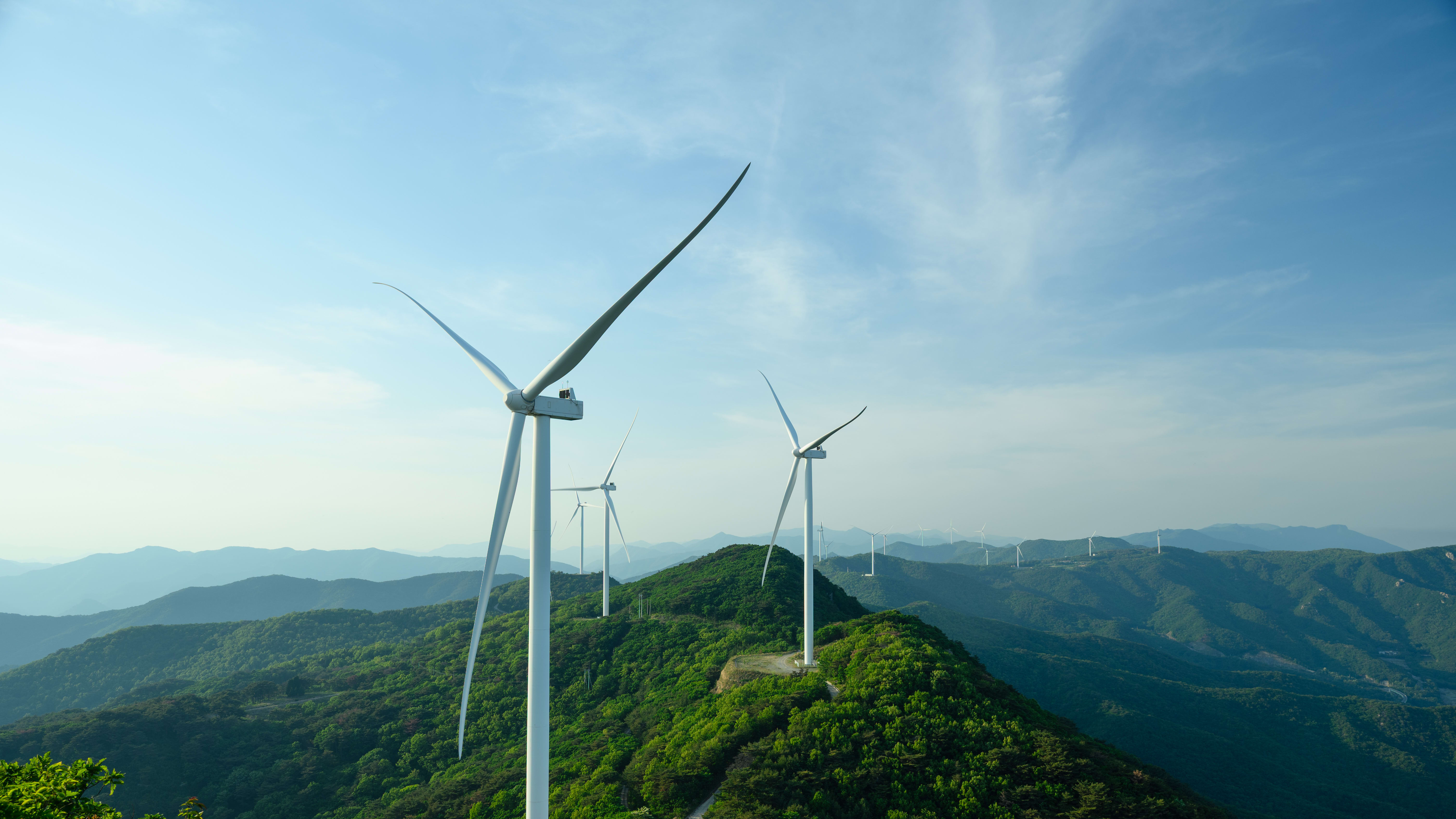 Multiple wind turbines on green forested mountain ridges under a blue sky