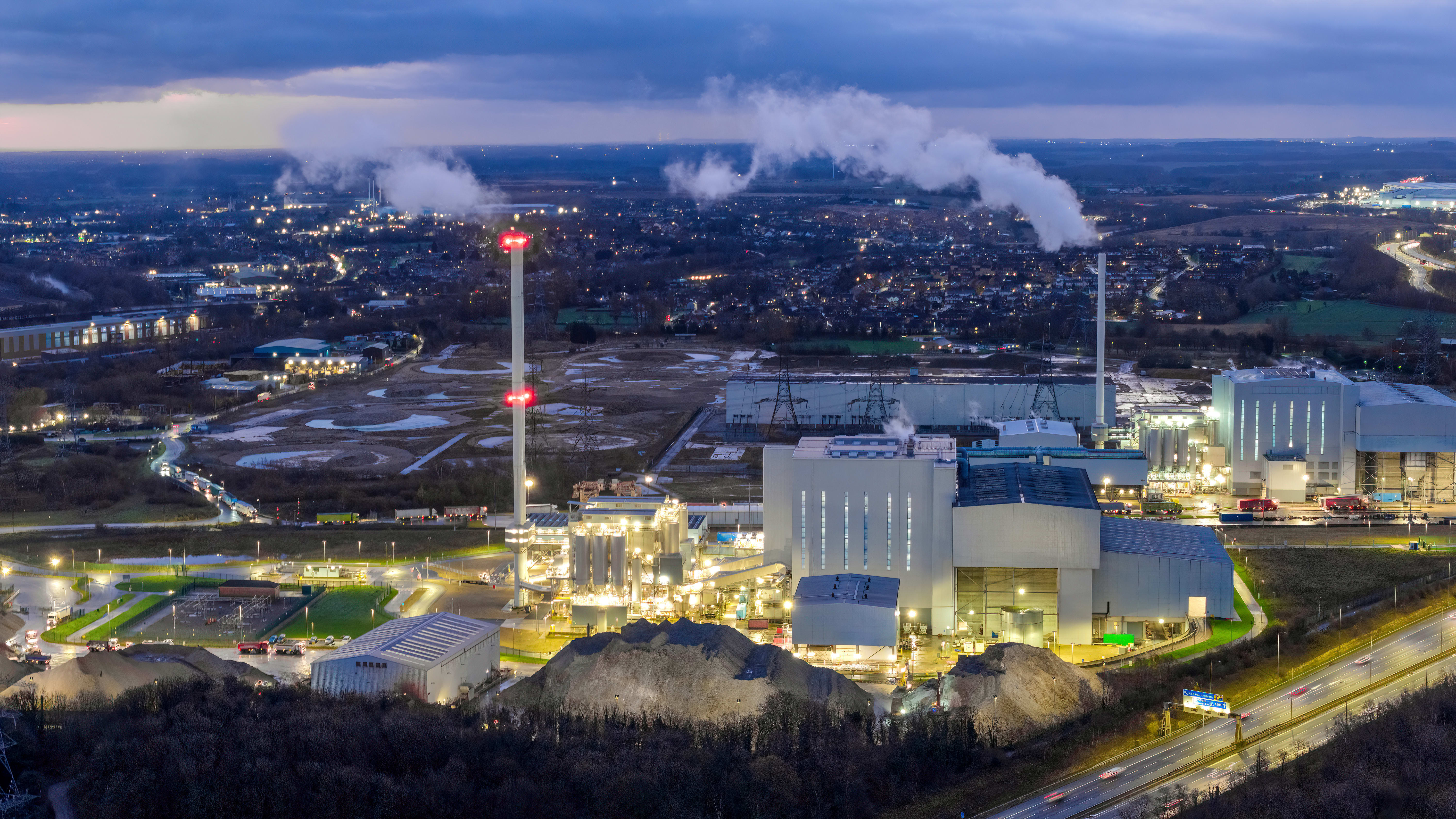 Aerial dusk view of an illuminated industrial energy plant with smoke stacks beside a motorway